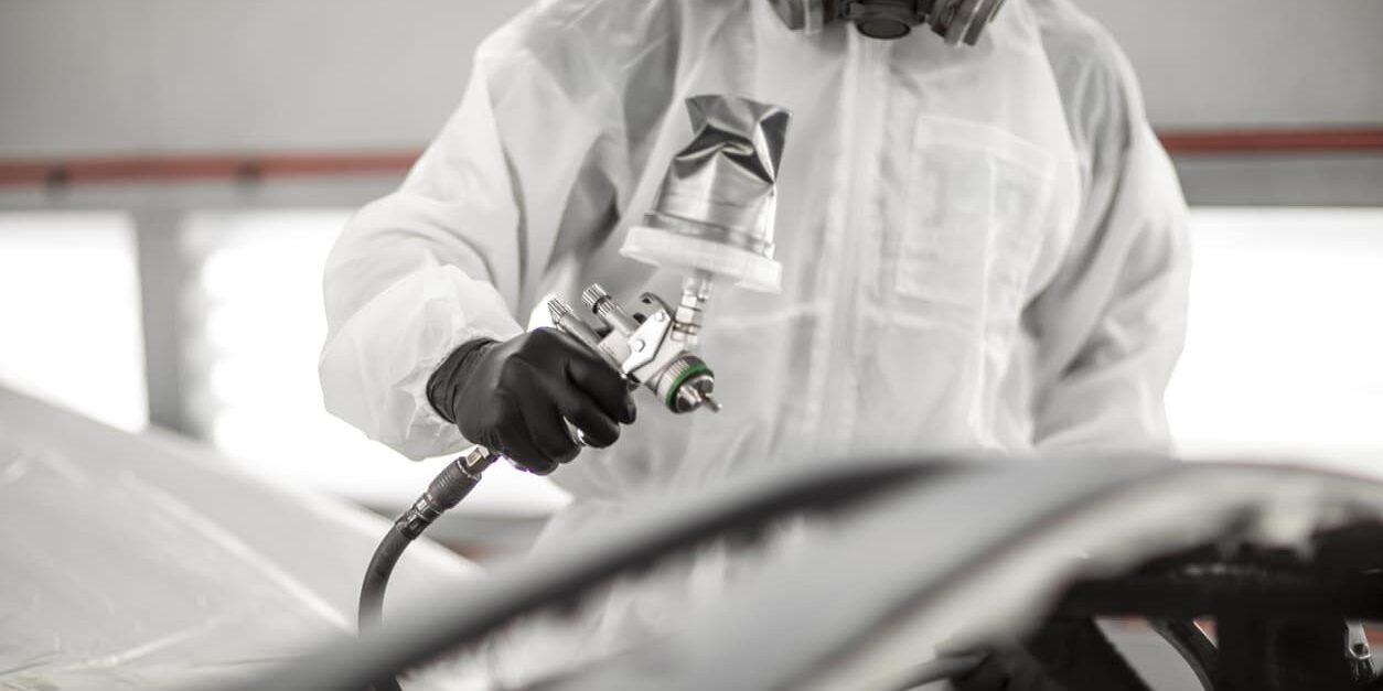 Car painter spraying a car part with a pressurized spray gun in a painting chamber.