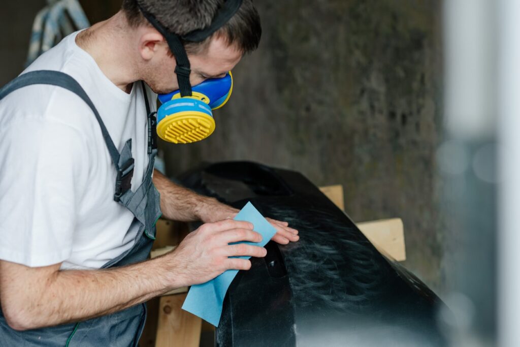 Young man sanding a car bumper with sandpaper. First step before painting car's plastic bumper cover. Worker using protective gear while working to keep from inhaling harmful fumes.