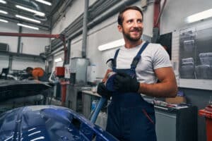 Smiling auto mechanic standing next to blue car body part and holding grinder while taking off sandpaper circle from it