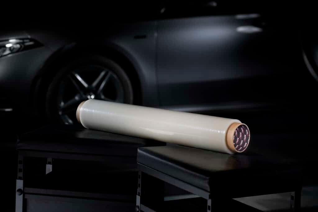 A large roll of film rests on a black table, with a sleek, dark car in the background in a dimly-lit environment.