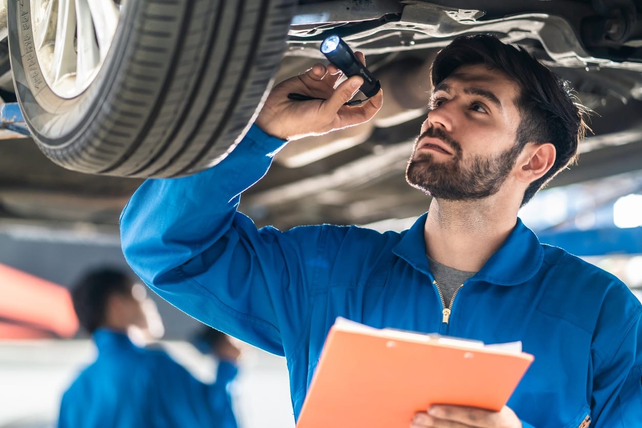Vehicle service maintenance handsome man checking under car condition in garage. Automotive mechanic pointing flash light on wheel following maintenance checklist document.