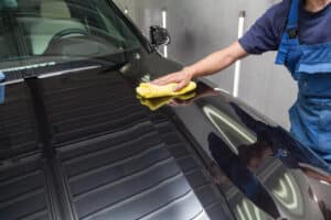 A male worker in blue overalls wipes the hood of a black car with a yellow microfiber cloth during polishing before applying a protective nano ceramic coating. Auto service industry.