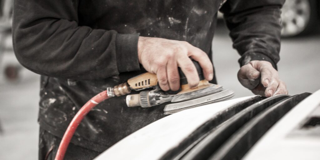 Body repair technician removing excess filling from an accident-damaged car bumper using a pneumatic sander.