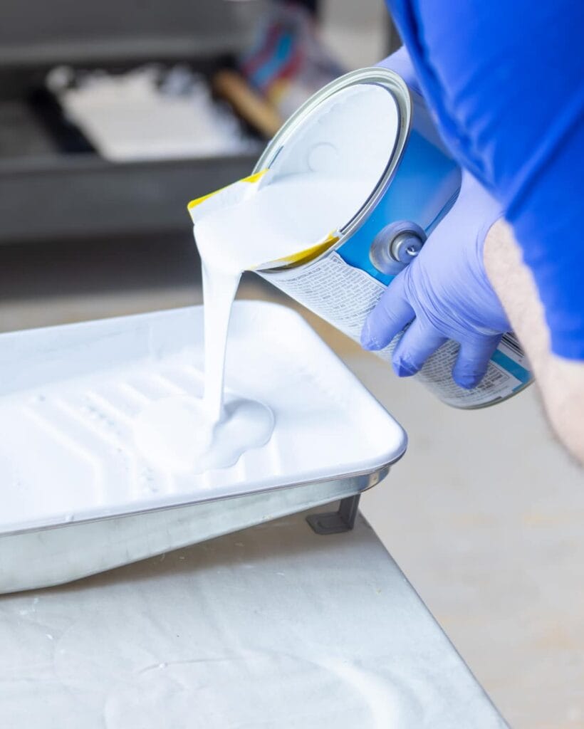 A person wearing blue gloves pours white paint from a can into a metal paint tray