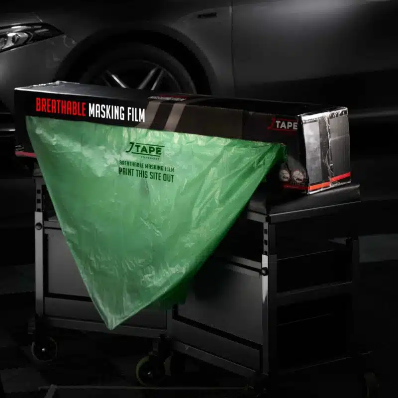 A green masking film is pulled from the box in a triangular shape and drapes over a black stand in front of a dark-coloured car.