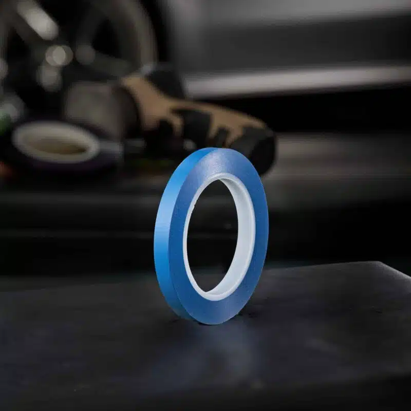 A roll of blue tape stands upright on a dark surface, with blurred vehicle parts in the background.