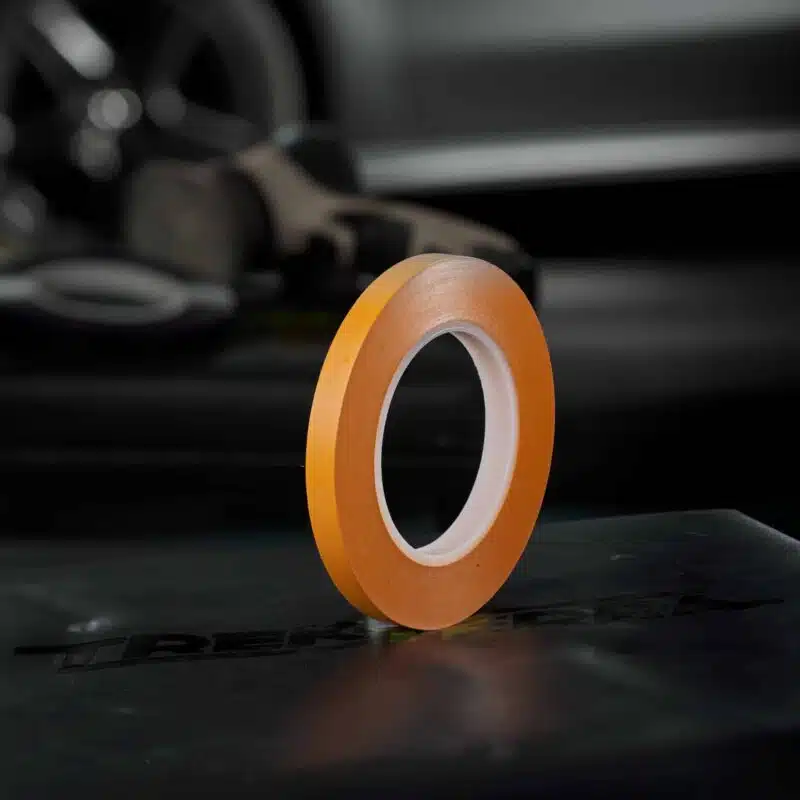 A roll of JTAPE's thin Orange Fine Line Tape rests on a smooth, dark surface, with blurred tape and industrial equipment visible in the dark background.
