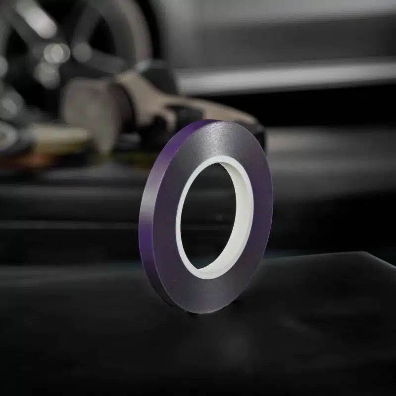 A roll of purple tape stands upright on a dark surface, with blurred vehicle parts in the background