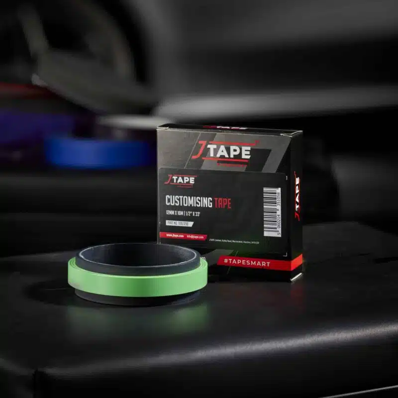 A roll of bright green tape lies flat on a dark surface next to a black box labelled "JTAPE Customising Tape". Blurred vehicle parts are in the background.