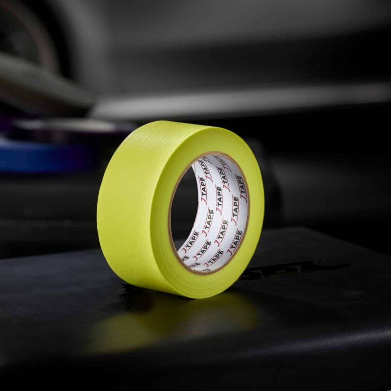 A roll of bright yellow wide tape, standing upright on a dark surface, with a blurred background that includes another roll of tape and a car door.