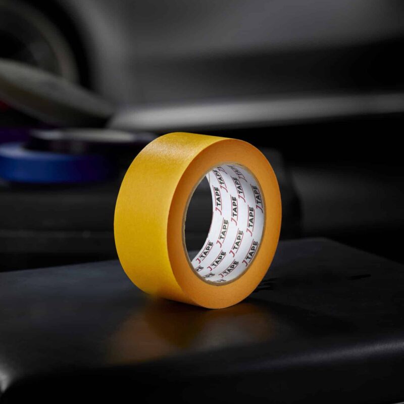 A roll of bright orange wide tape, standing upright on a dark surface, with a blurred background that includes another roll of tape and a car door.