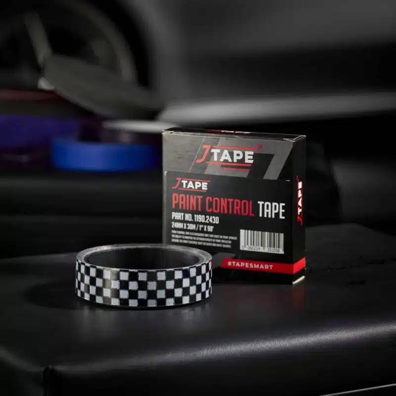 A roll of black and white checkered tape lies flat on a dark surface next to a black box labelled "JTAPE Paint Control Tape". Blurred vehicle parts are in the background.