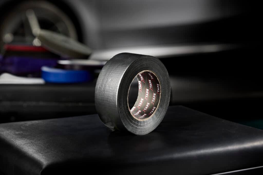 A roll of black masking tape sits on a dark surface, with a blurred background suggesting an industrial or workshop setting