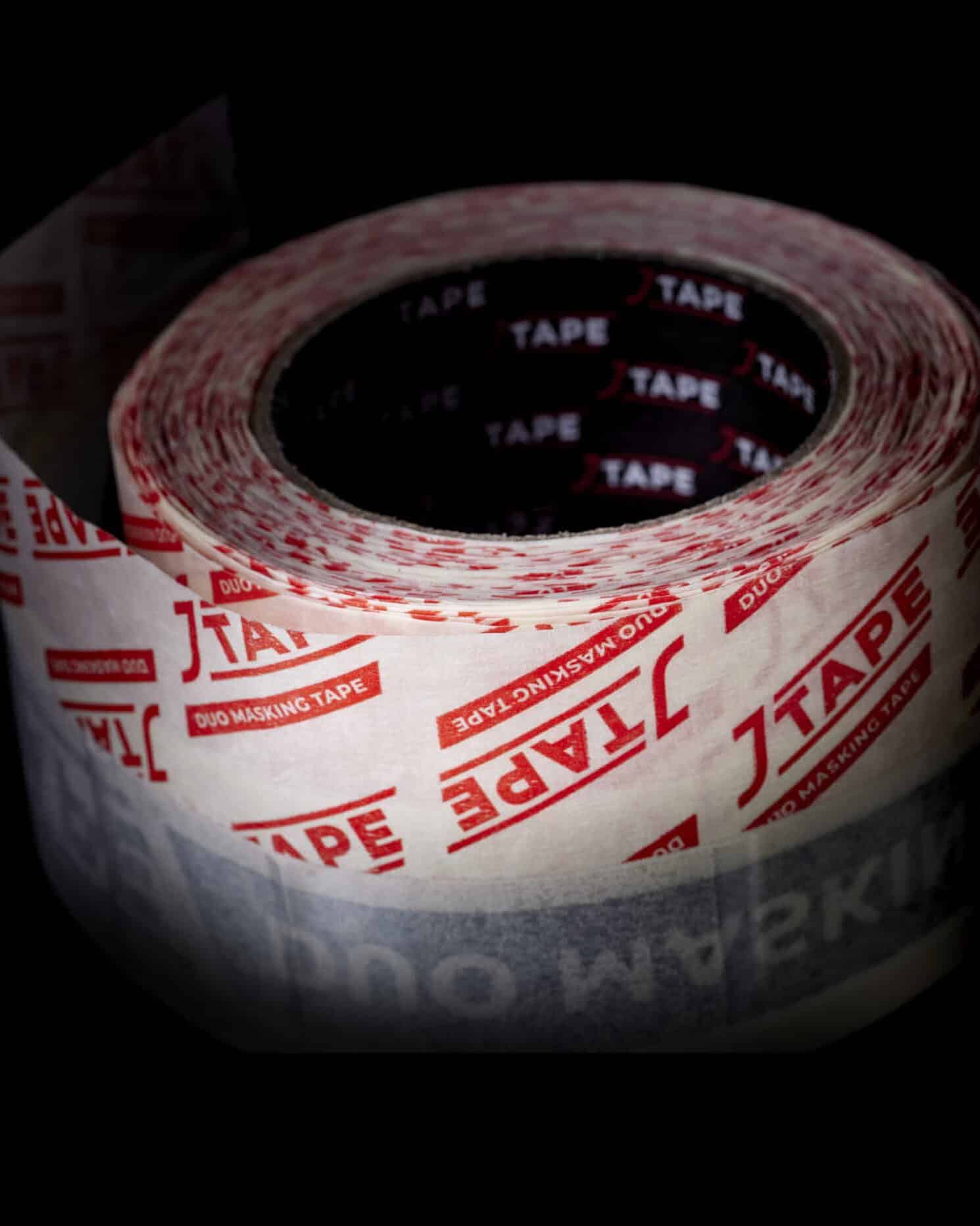 Close up of a slightly unrolled white and red patterned tape labelled 'JTAPE Duo Masking Tape' lay on a black surface.
