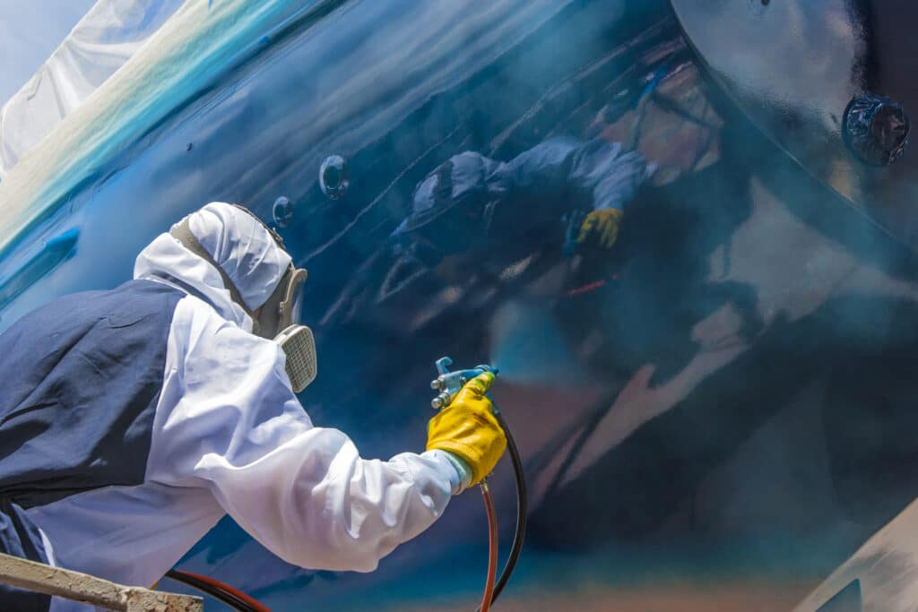 worker in full body protection suit and mask is spray painting the hull of a sailing yacht as final work of corrosion protection