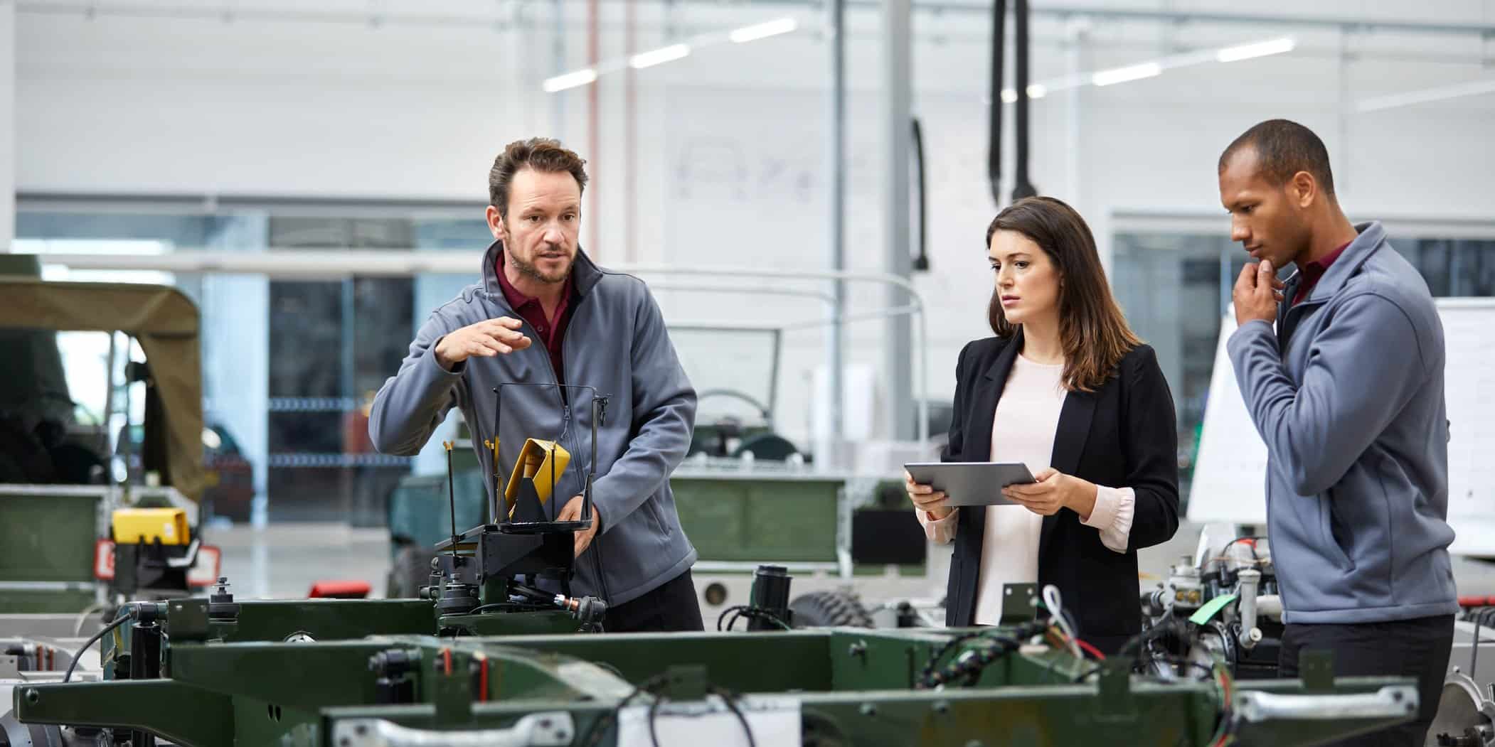 three people in a modern factory setting look intently at machinery. The woman holds a tablet.