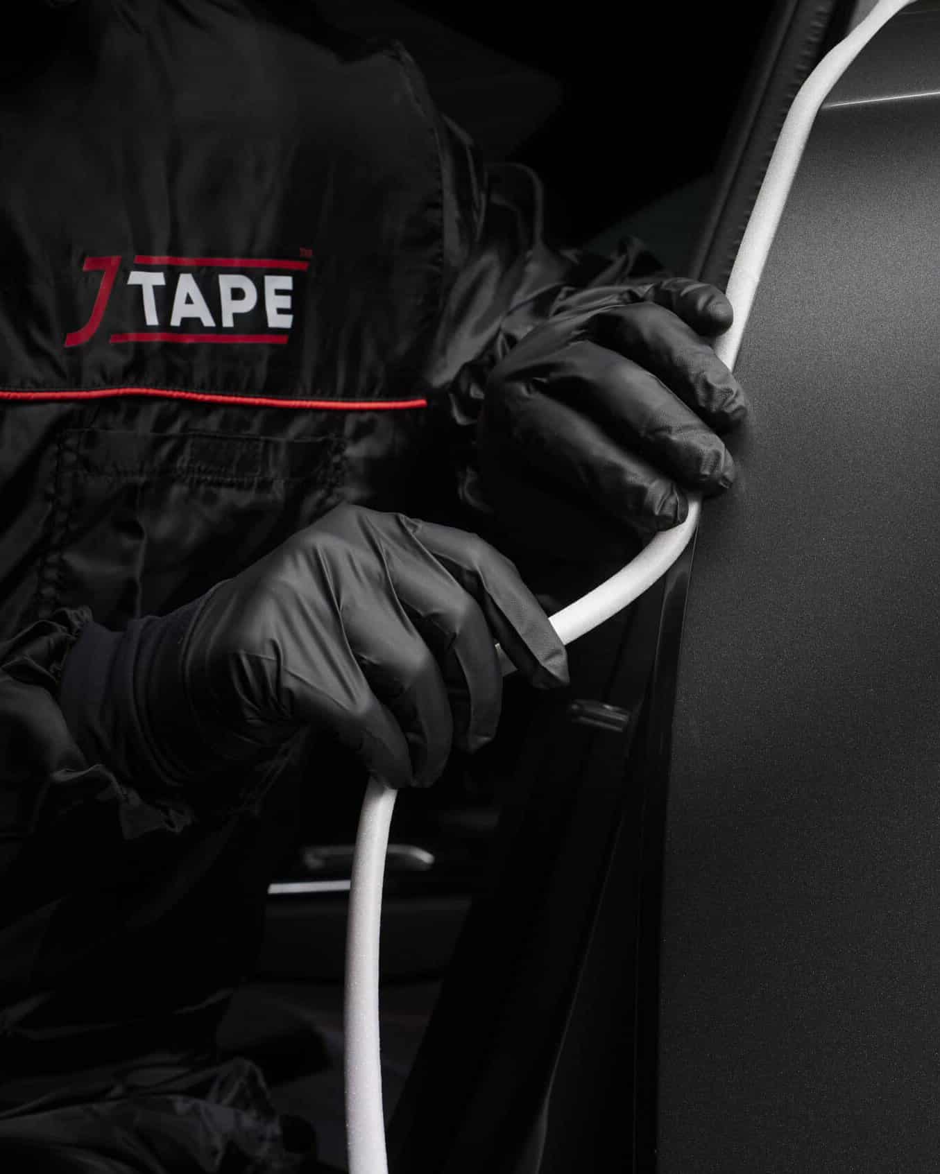 A person in black overalls with the JTAPe logo on, and black gloves applying white thin foam tape to an edge on a car.