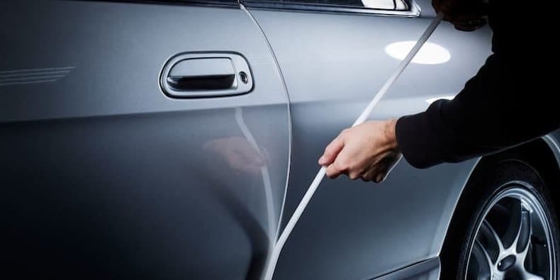 A person pulls white foam tape away from a silver car door edge, peeling the tape off. The car door handle and rear wheel are visible.