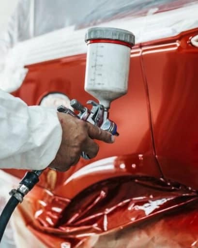 A worker in protective gear spray-paints a car in vibrant red inside a spray booth