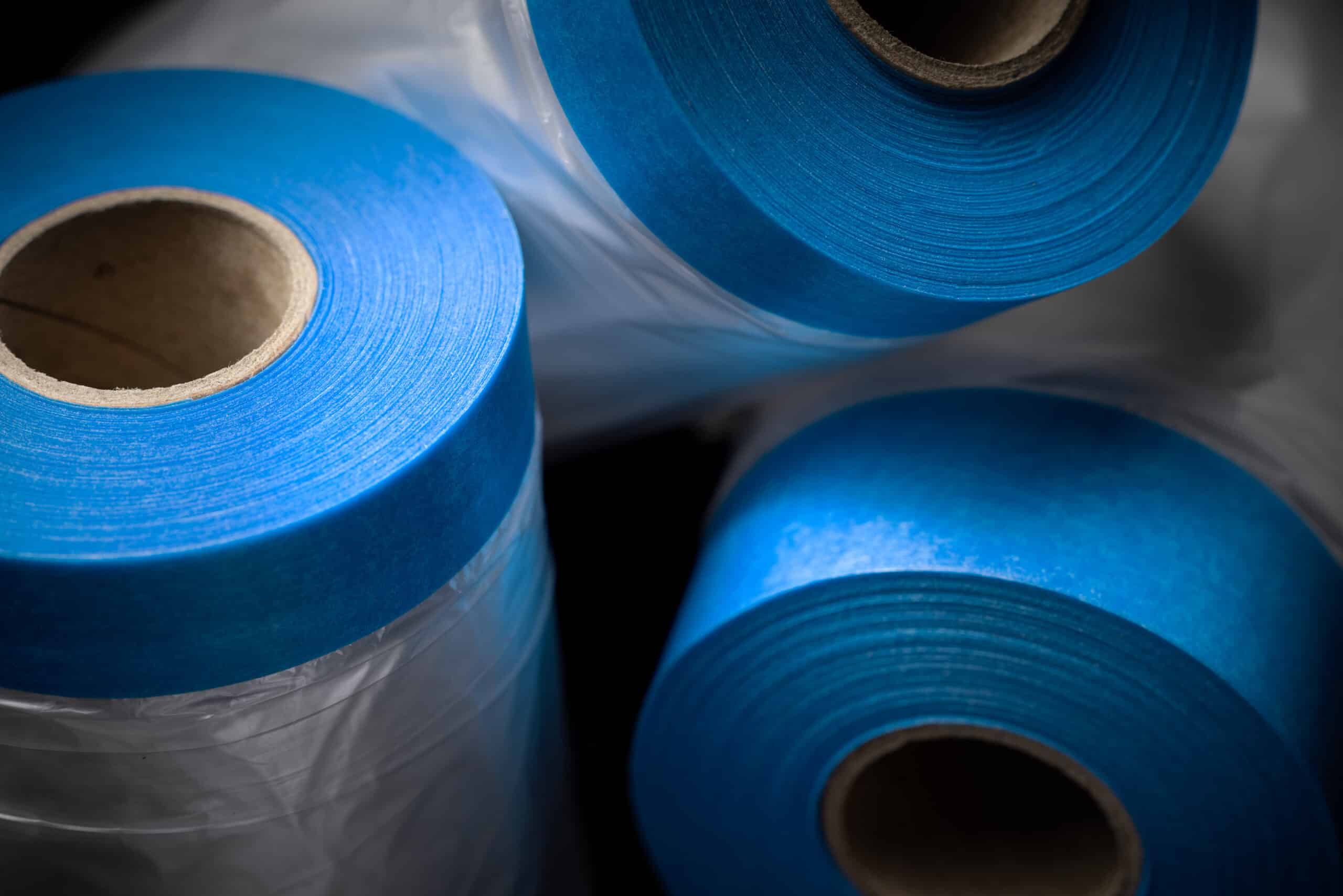 Three tightly wound rolls of bright blue film, wrapped in protective plastic, are positioned against a dark background.