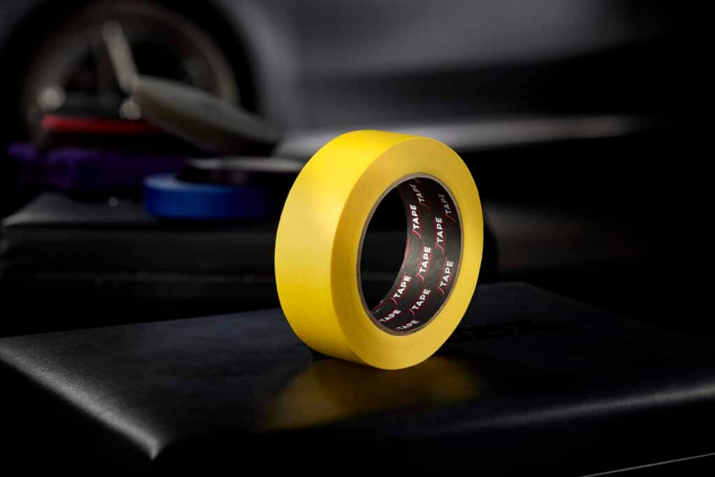 A roll of yellow tape sits on a dark surface, with a blurred background suggesting an industrial or workshop setting