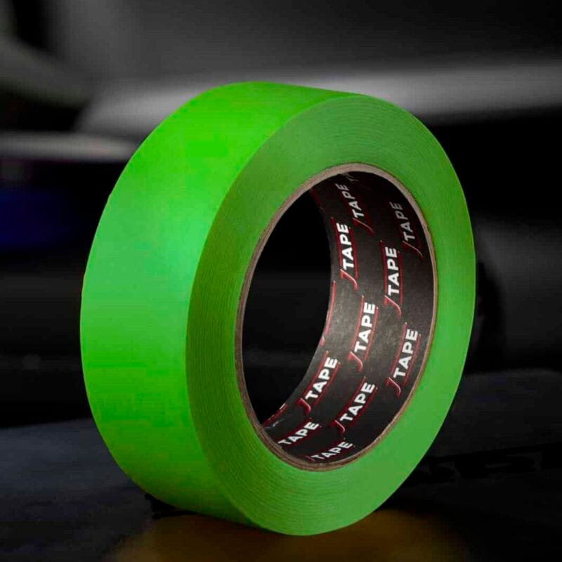 A roll of bright green tape, standing upright on a dark surface, with a blurred background that includes another roll of tape and a car door.