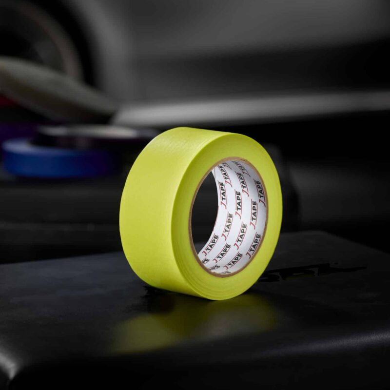 A roll of 120ºC Lime Precision Masking Tape rests on a smooth, dark surface, with a blurred tapes and a dark car in the background.